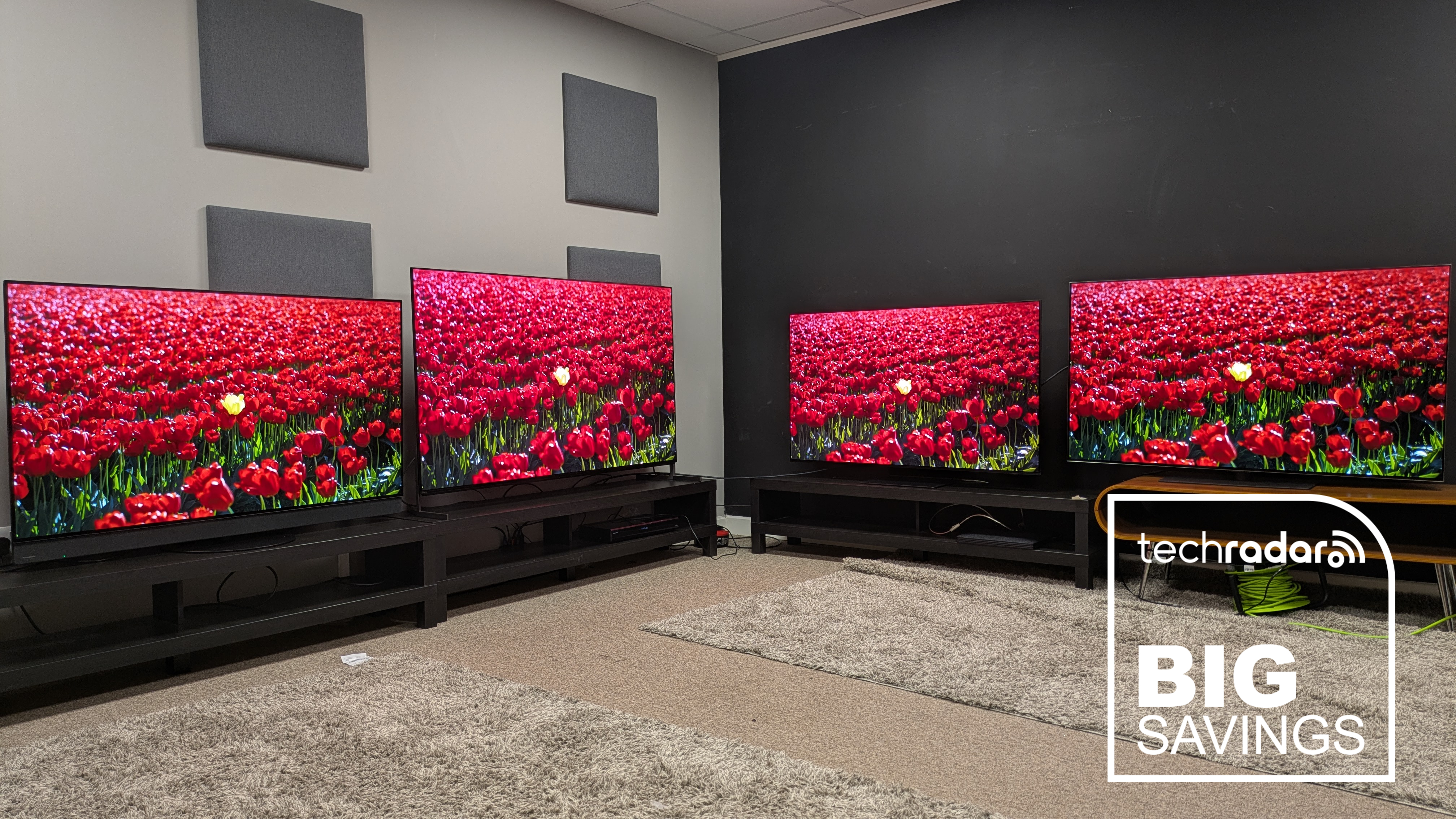 Deal image with four OLED TVs displaying red flowers on their screens with big savings logo in white