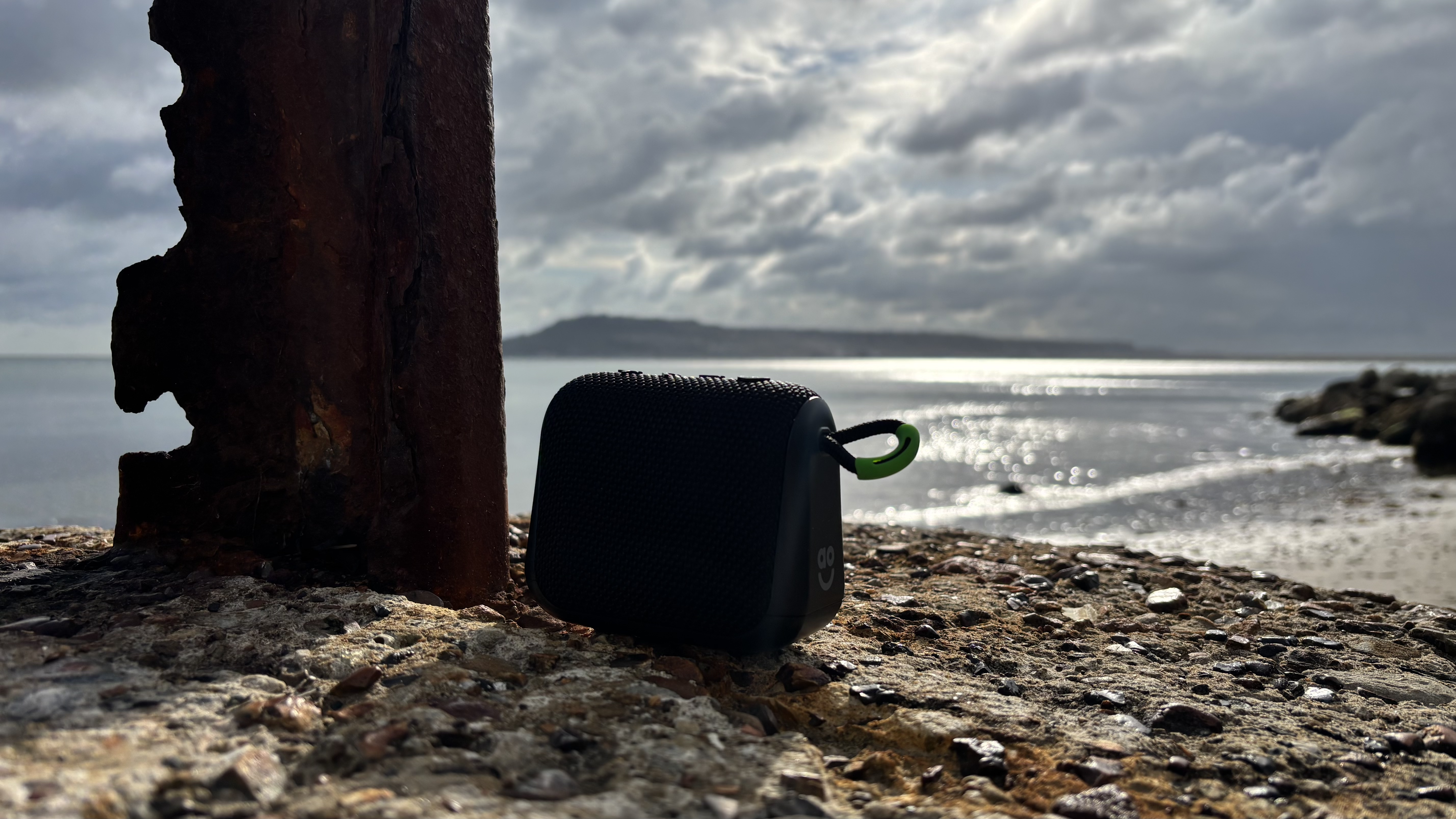 Black AO mini portable Bluetooth speaker perched on a rock with a harbor and island in the background